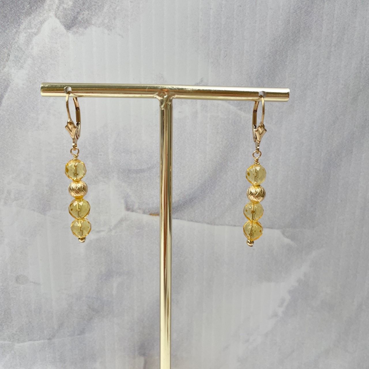 Aan een 14K gold filled brissure oorbel, dit is een sluitbaar oorbelhaakje, met leliebloem, hangen drie citrien kralen met ertussen een bewerkte gold filled kraal. De citrien kralen zijn van AA kwaliteit en hebben heel veel facetjes. De oorbellen hangen aan een standaardje.