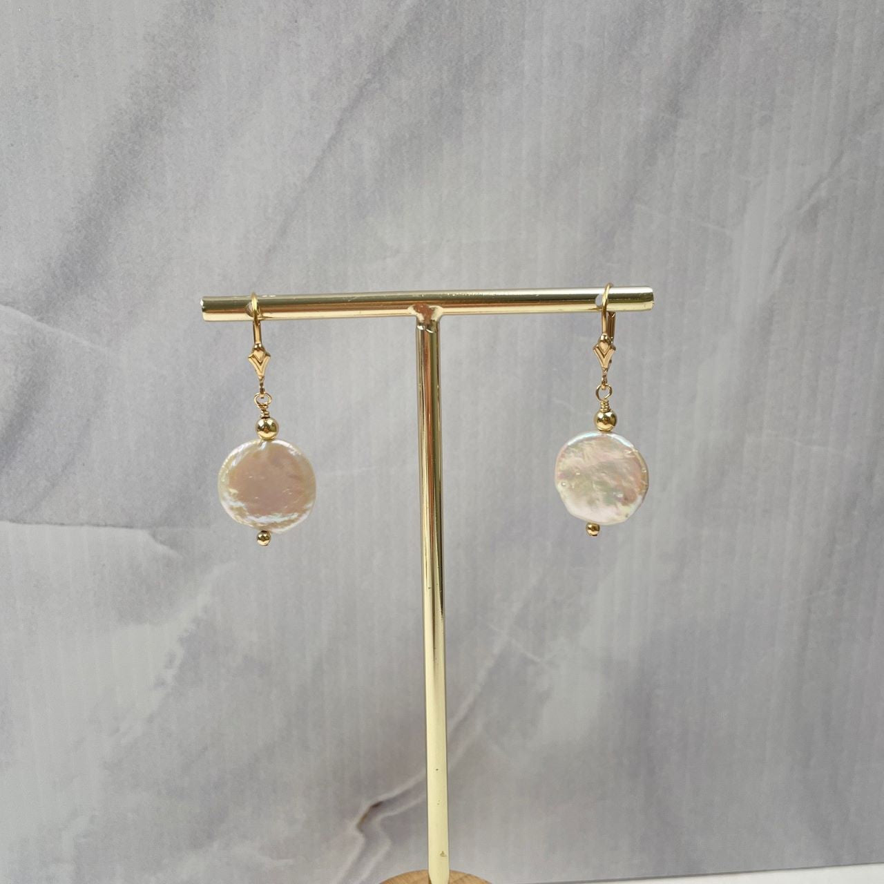 Aan een 14K gold filled brissure met lelie, hangen, tussen twee gold filled kralen, zogeheten zoetwaterparel coins of munten. Hier hangen de oorbellen aan een standaard.